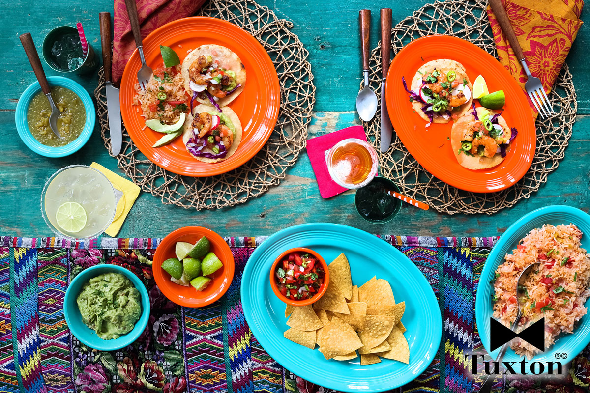 Colorful Mexican meal on a vibrant table with tacos, chips and salsa, guacamole, rice, lime wedges, drinks, and sauces on patterned plates and bowls. The Tuxton logo is in the bottom right corner.