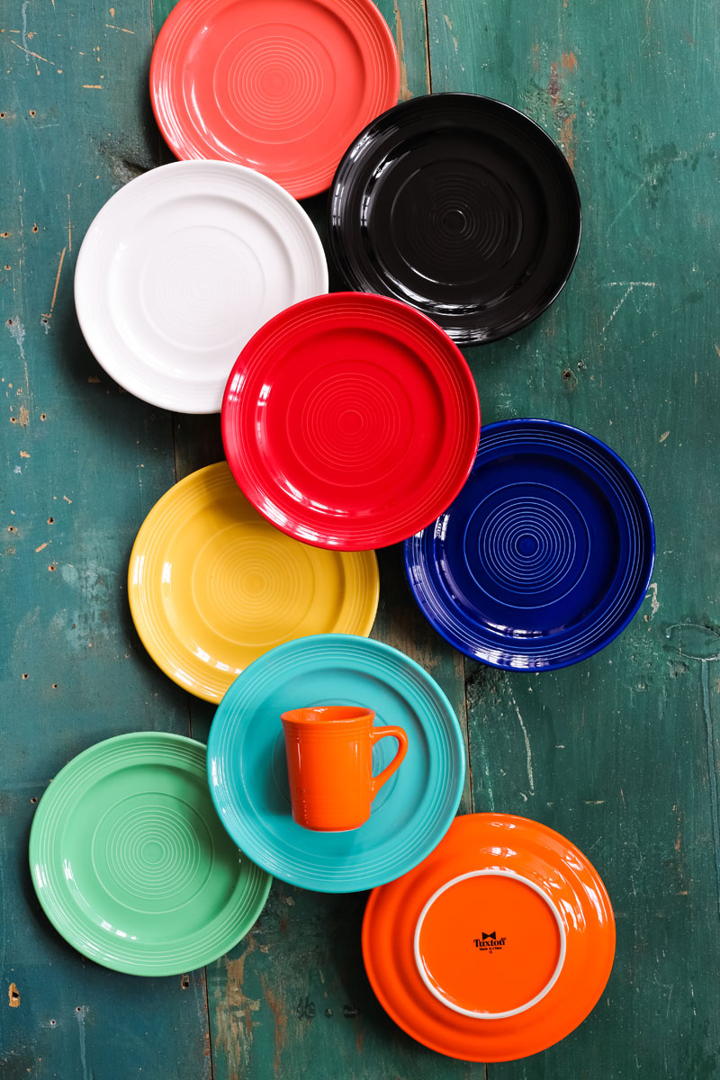 Ten colorful plates (red, black, white, blue, yellow, orange, green, turquoise) are arranged in a loose spiral on a distressed teal wooden surface, with an orange cup placed on one turquoise plate.
