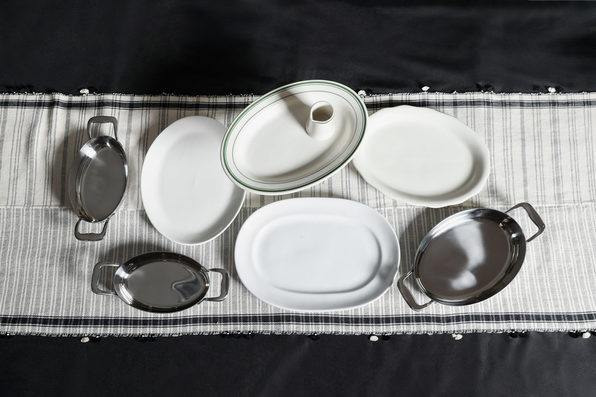 Seven oval serving platters, including metal and white ceramic dishes, are arranged on a striped table runner over a black tablecloth.