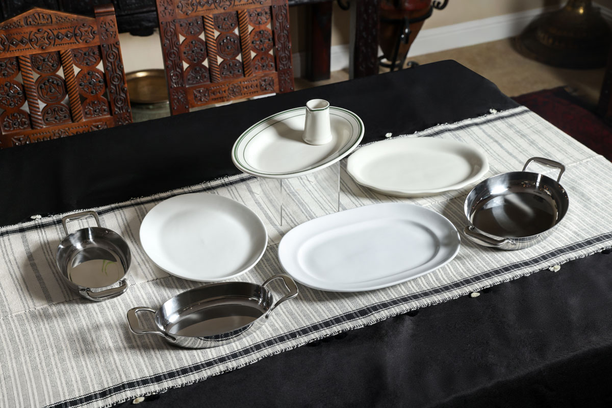 Six empty serving dishes, including three stainless steel bowls with handles, two white oval platters, and one oval platter with a cup in the center, are arranged on a black and white tablecloth over a dark wooden table.