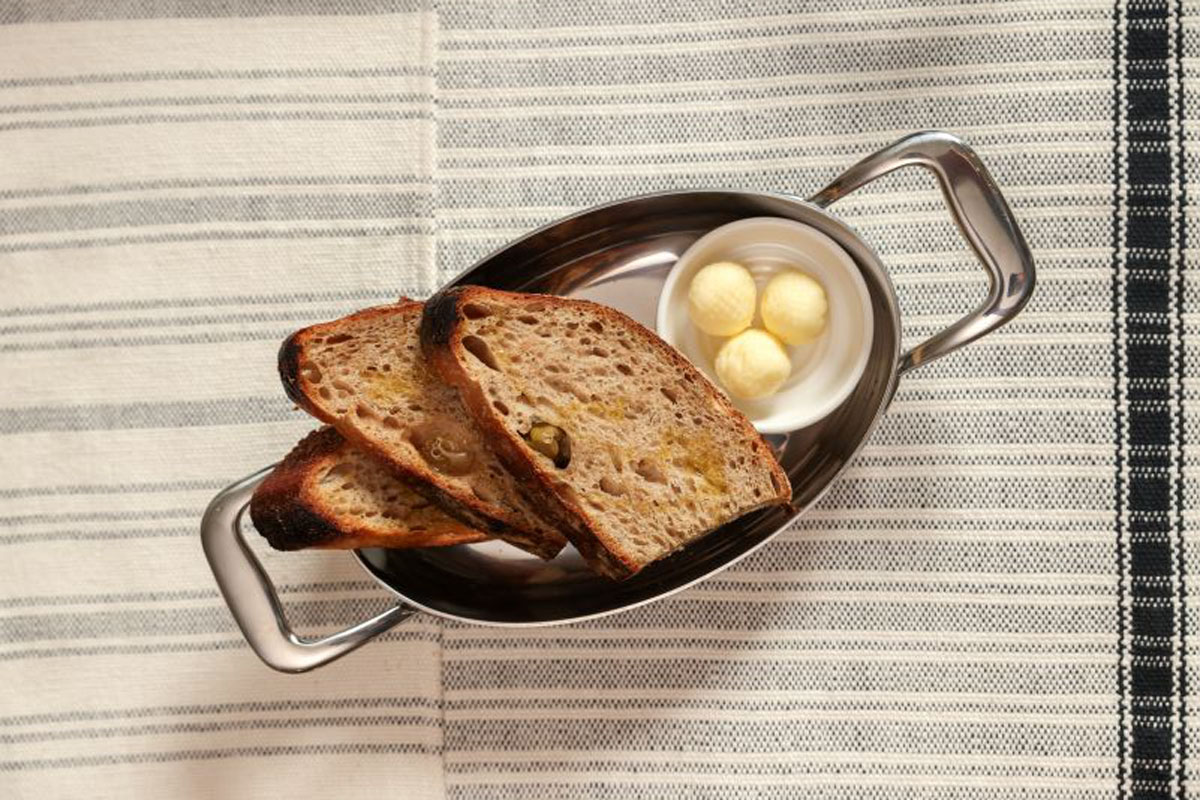 Two slices of toasted bread and three small balls of butter are served in a metal dish with handles, placed on a striped fabric tablecloth.