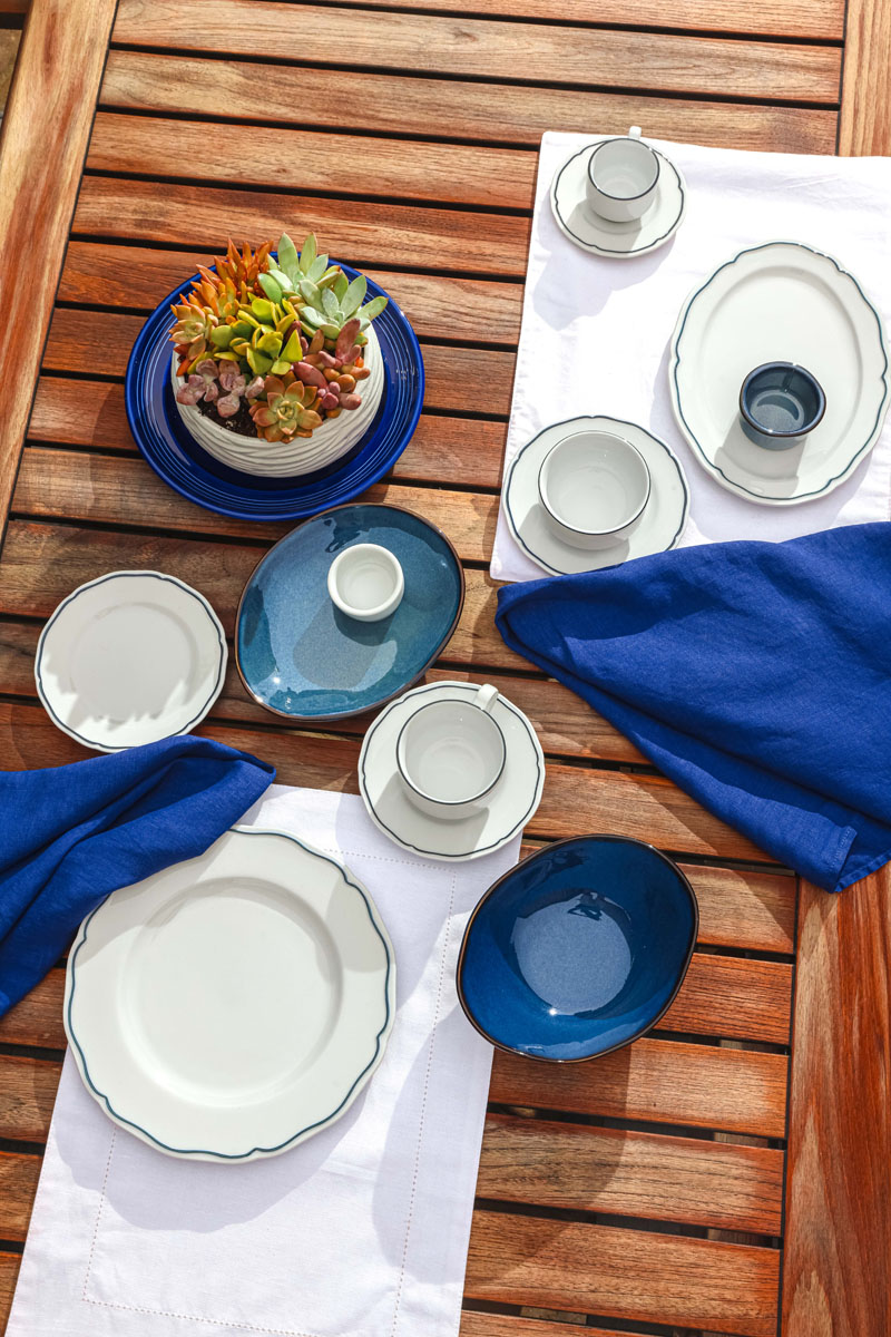 A wooden table set with white and blue dishes, cups, and bowls, two royal blue napkins, white placemats, and a centerpiece of colorful succulents in a bowl.
