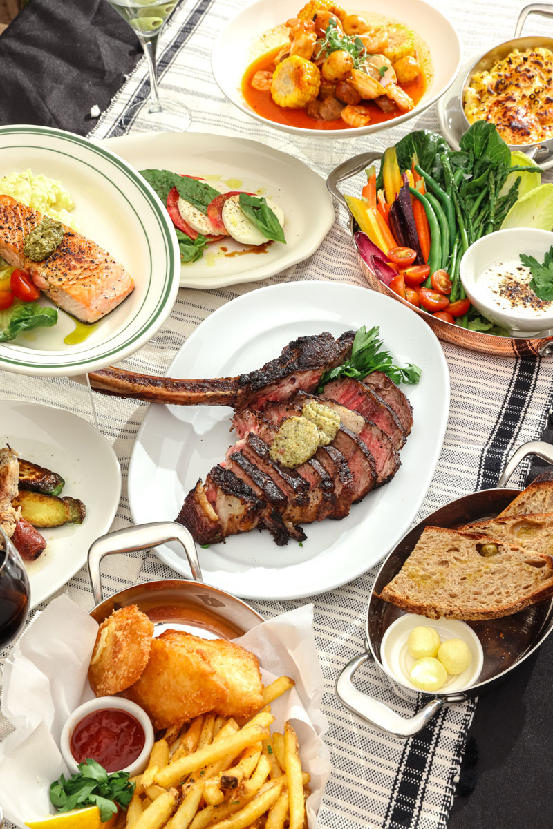 A variety of gourmet dishes on a table, including grilled steak, salmon, shrimp, fresh vegetables, gratin, bread with butter, fish and chips with fries, and assorted small plates.