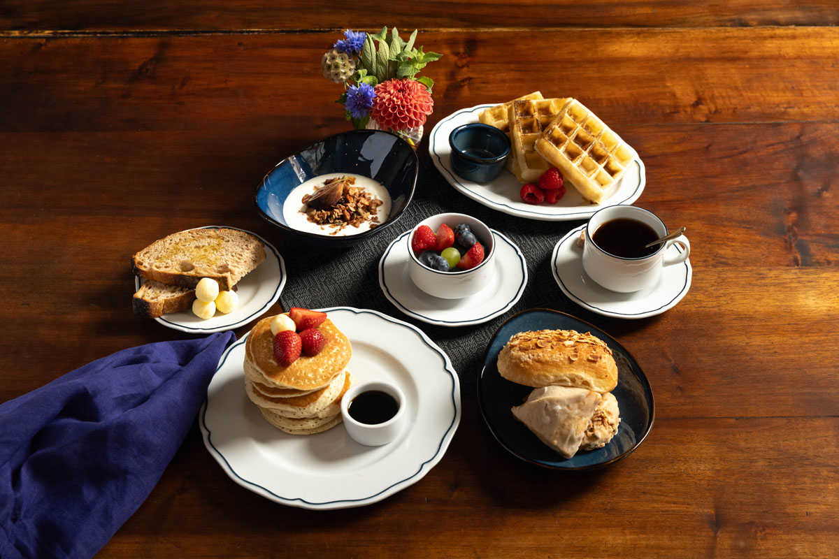 A wooden table set with pancakes topped with strawberries, waffles with raspberries and syrup, toast with butter, mixed fruit, yogurt with granola, a scone, two cups of coffee, and a small flower arrangement.