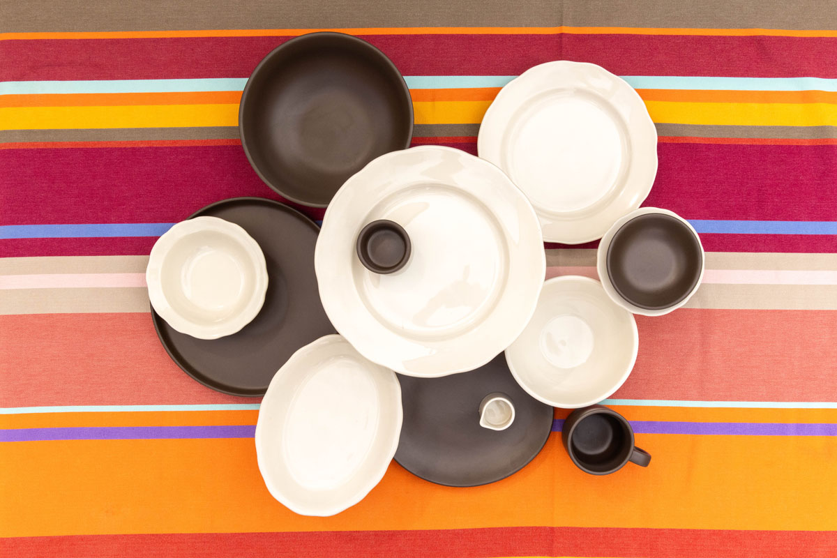 A top view of assorted white and dark brown ceramic plates, bowls, and cups arranged on a colorful striped tablecloth with red, orange, yellow, and light blue stripes.