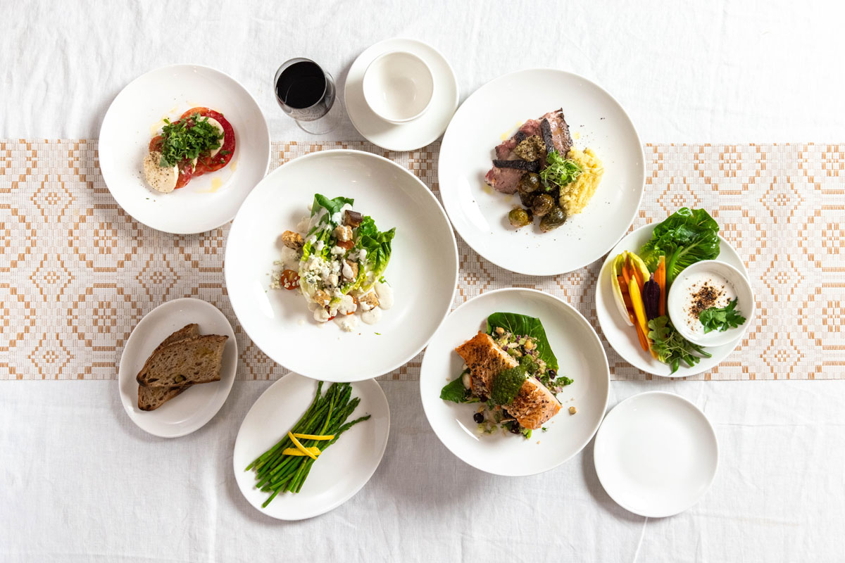 A top view of a white tablecloth with several plates of food, including salad, grilled fish, meat with potatoes, sliced bread, green beans, a glass of red wine, and an empty bowl and plate.