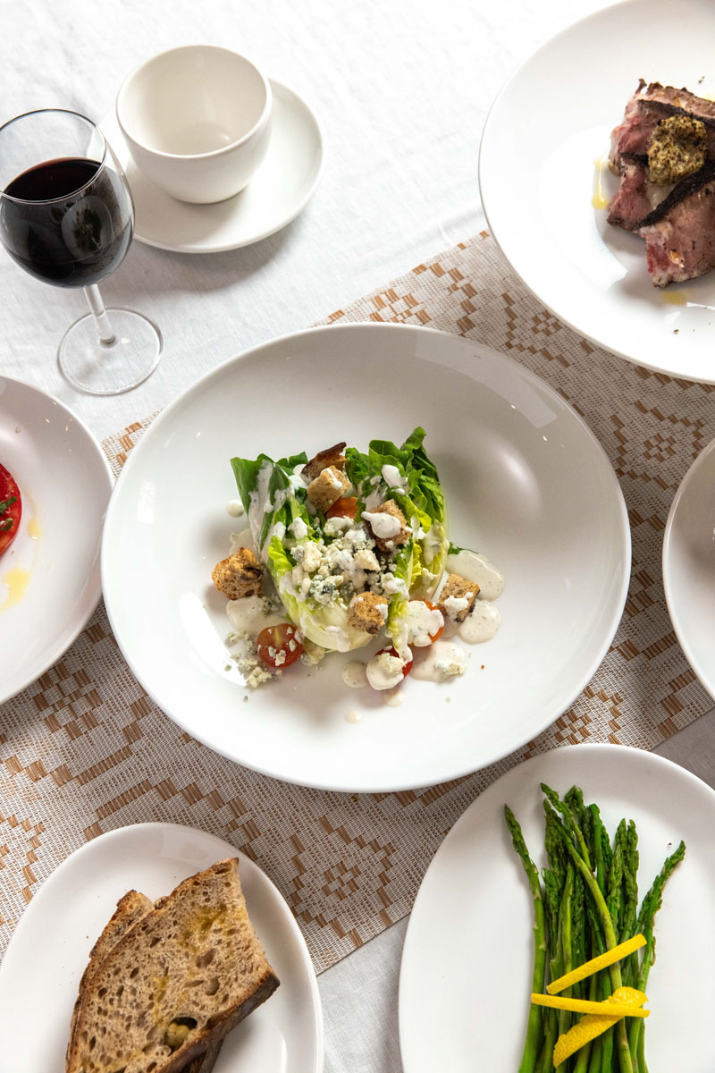 A table set with various dishes, including a wedge salad with blue cheese, tomatoes, and croutons, grilled asparagus, sliced bread, a plate of roasted meat, a glass of red wine, and white dishware on a white cloth.