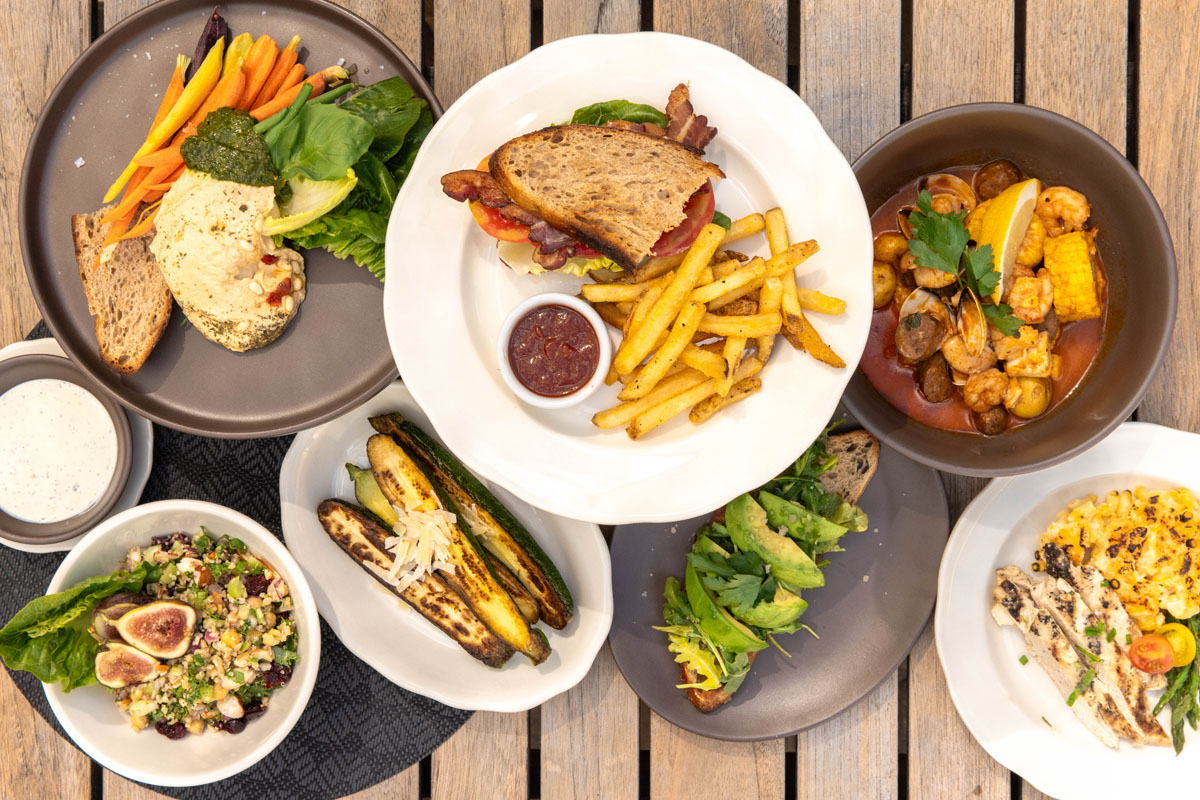An overhead view of various dishes on a wooden table, including a sandwich with fries, roasted vegetables, salad, soup with bread, pasta, and assorted fresh vegetables.