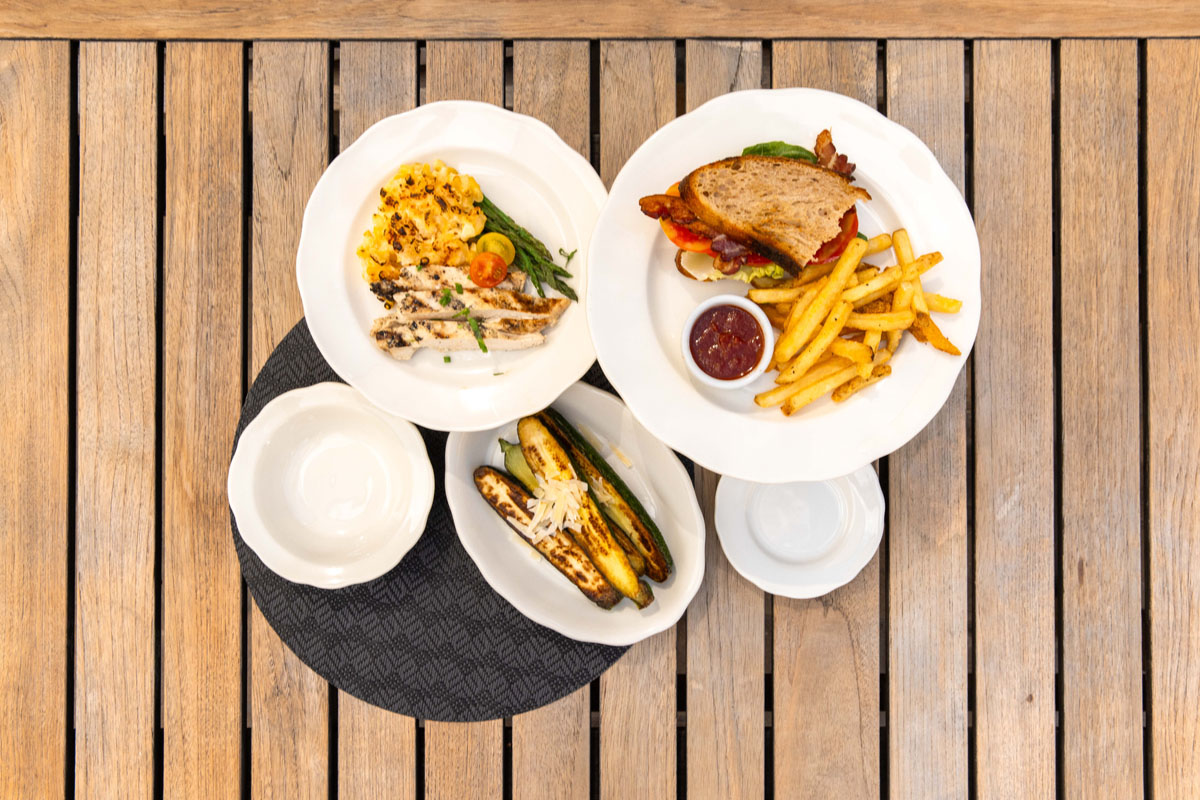 Three plates on a wooden table: one with grilled chicken and rice, one with a sandwich, fries, and ketchup, and one with grilled zucchini, plus two small empty dishes nearby.
