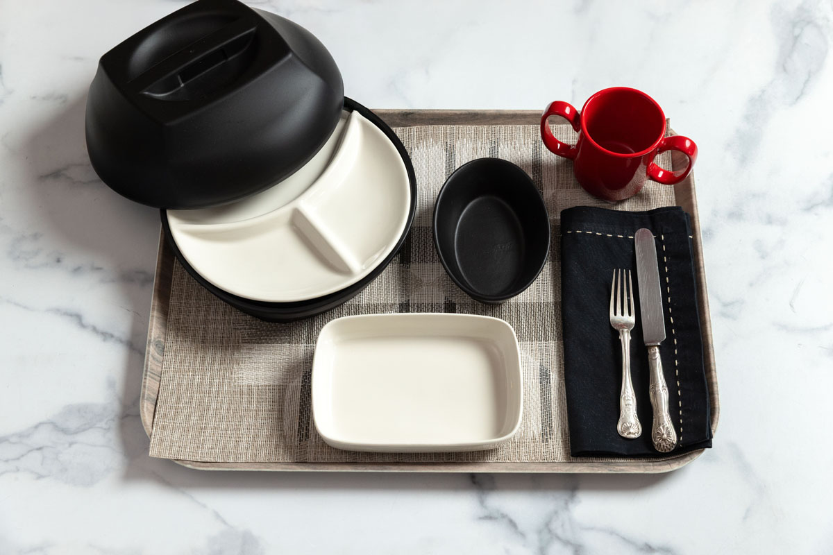 A tray with a divided plate covered by a black lid, a rectangular white dish, a small black oval bowl, a red two-handled cup, a fork and knife on a black napkin, all on a marble surface.