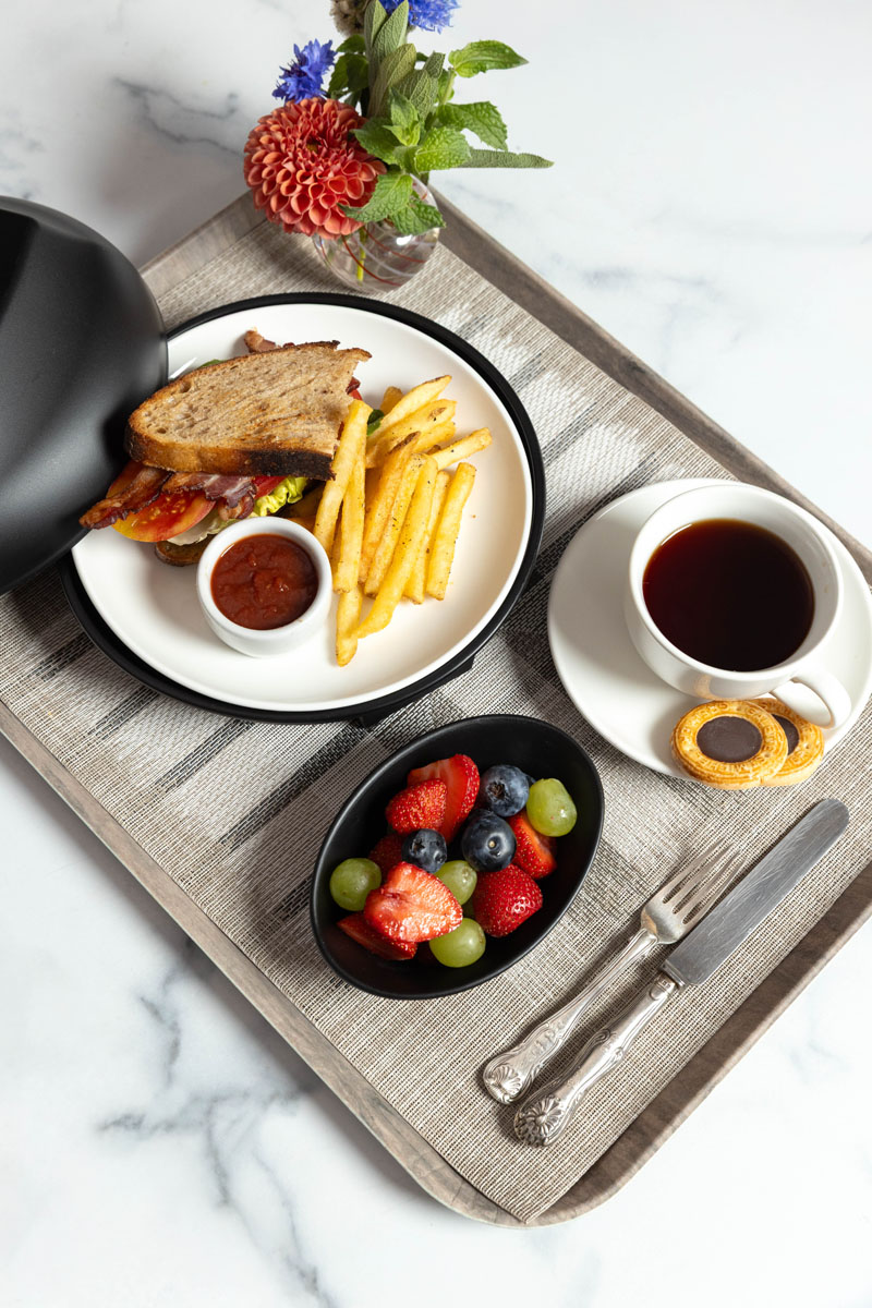 A breakfast tray with a BLT sandwich, French fries with ketchup, mixed fruit, a cup of black coffee with a cookie, a knife and fork, and a small vase with red and blue flowers on a gray placemat.