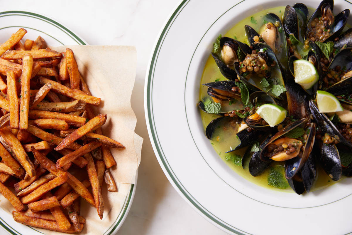 A plate of steamed mussels in broth with lime wedges and herbs, served next to a basket of crispy French fries on white plates with green rims.