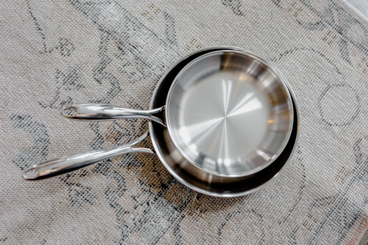 Two shiny stainless steel frying pans, one nested inside the other, placed on a textured, light-colored fabric surface.