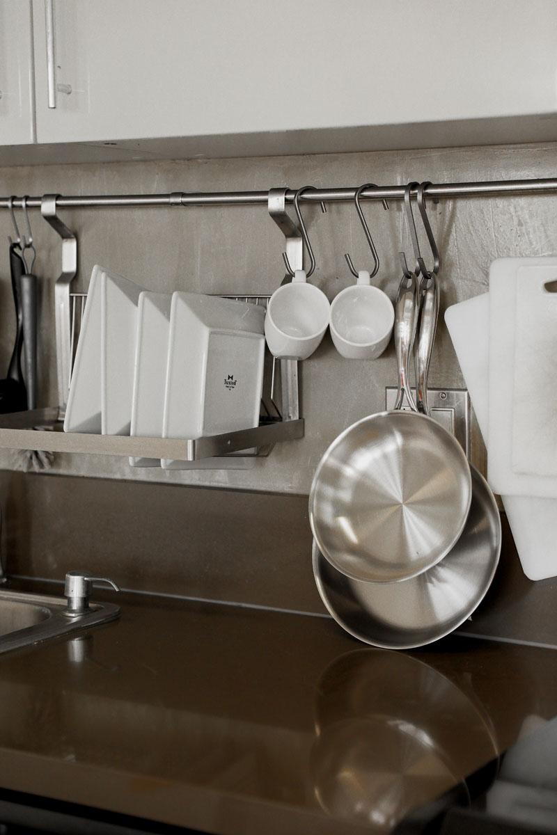 A modern kitchen with clean dishes in a drying rack, two white mugs hanging on hooks, stainless steel pans, utensils, and cutting boards organized neatly against a gray backsplash.