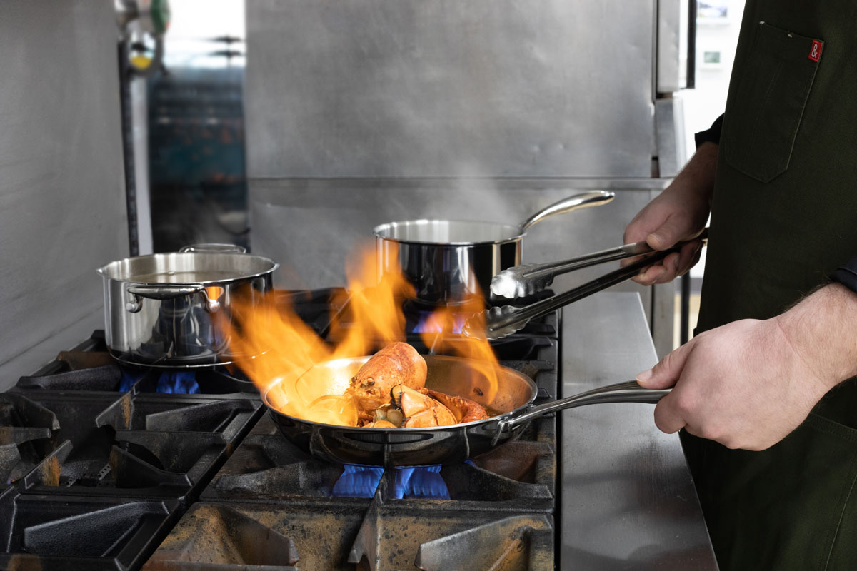 A cook uses tongs to sauté food in a flaming pan on a gas stove, with two pots boiling in the background in a commercial kitchen.