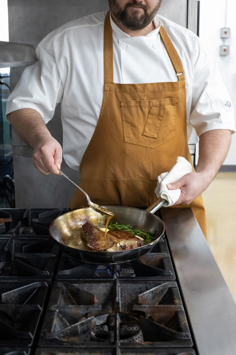 A chef in a white coat and brown apron is basting a steak with butter in a pan on a stovetop, holding a spoon in one hand and a towel in the other. Fresh herbs are also visible in the pan.