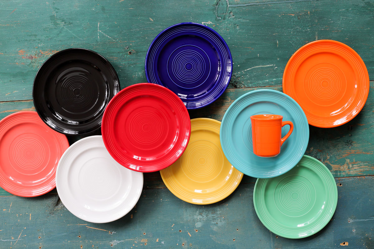 Nine colorful plates and one orange mug are arranged on a rustic green wooden surface. The plates are in various colors including black, blue, orange, red, pink, white, yellow, turquoise, and green.