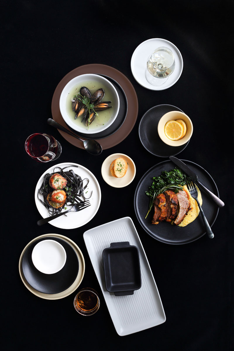 A top-down view of a table with assorted dishes, including mussels in broth, black pasta with seafood, roasted meat with greens, bread, lemon slices, and drinks on black and white plates.