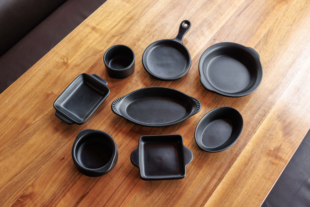 Eight black ceramic dishes of various shapes and sizes are arranged neatly on a wooden table, including round, oval, rectangular, and square pieces, as well as a small skillet with a handle.