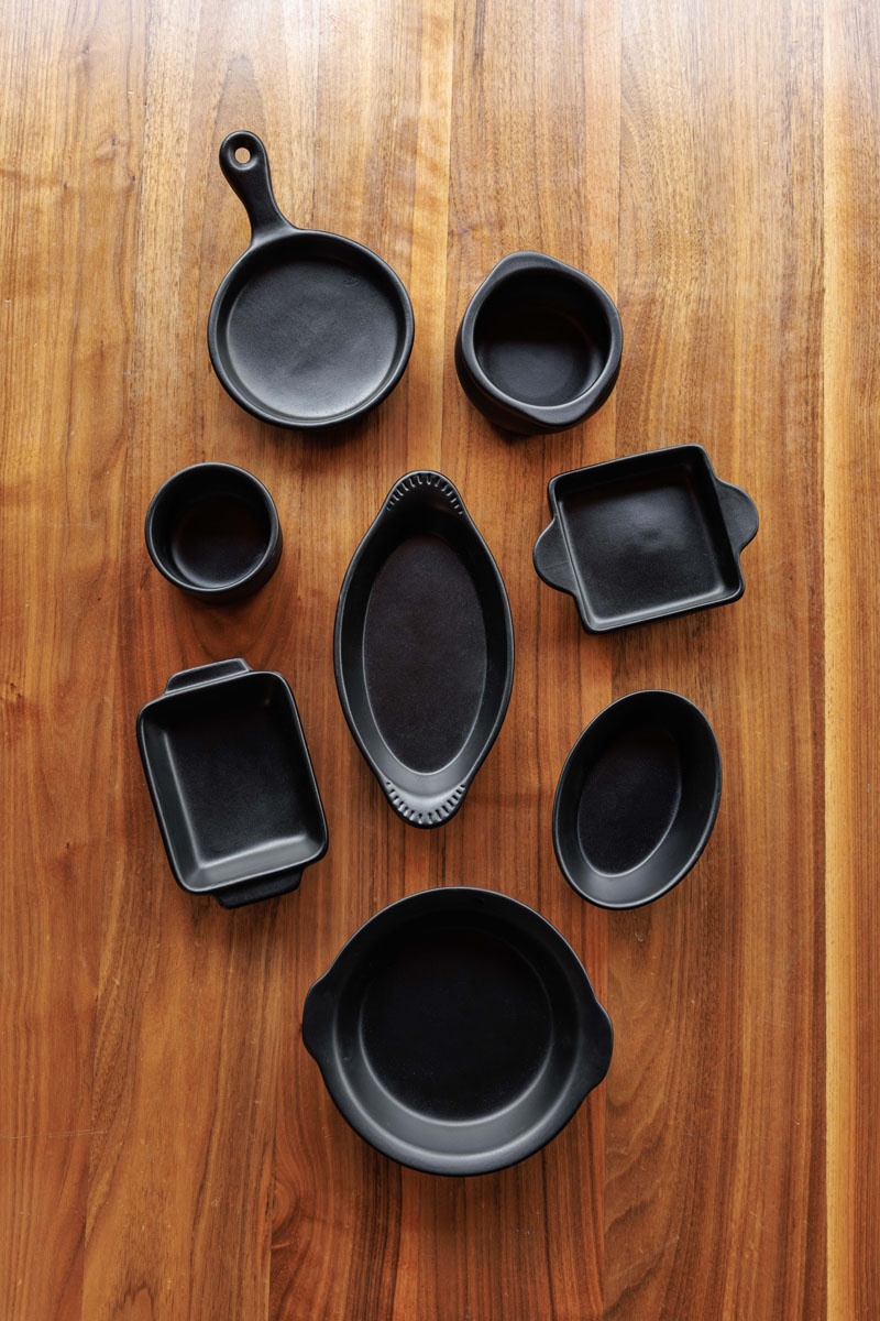 Eight black ceramic baking dishes of various shapes and sizes are arranged neatly on a wooden surface, viewed from above.