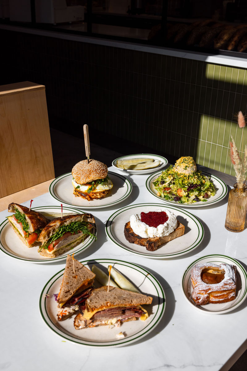 A table set with assorted dishes: sandwiches, a burger, a salad, toast with spread and fruit topping, and a pastry, all on white plates with green trim, in bright natural light.
