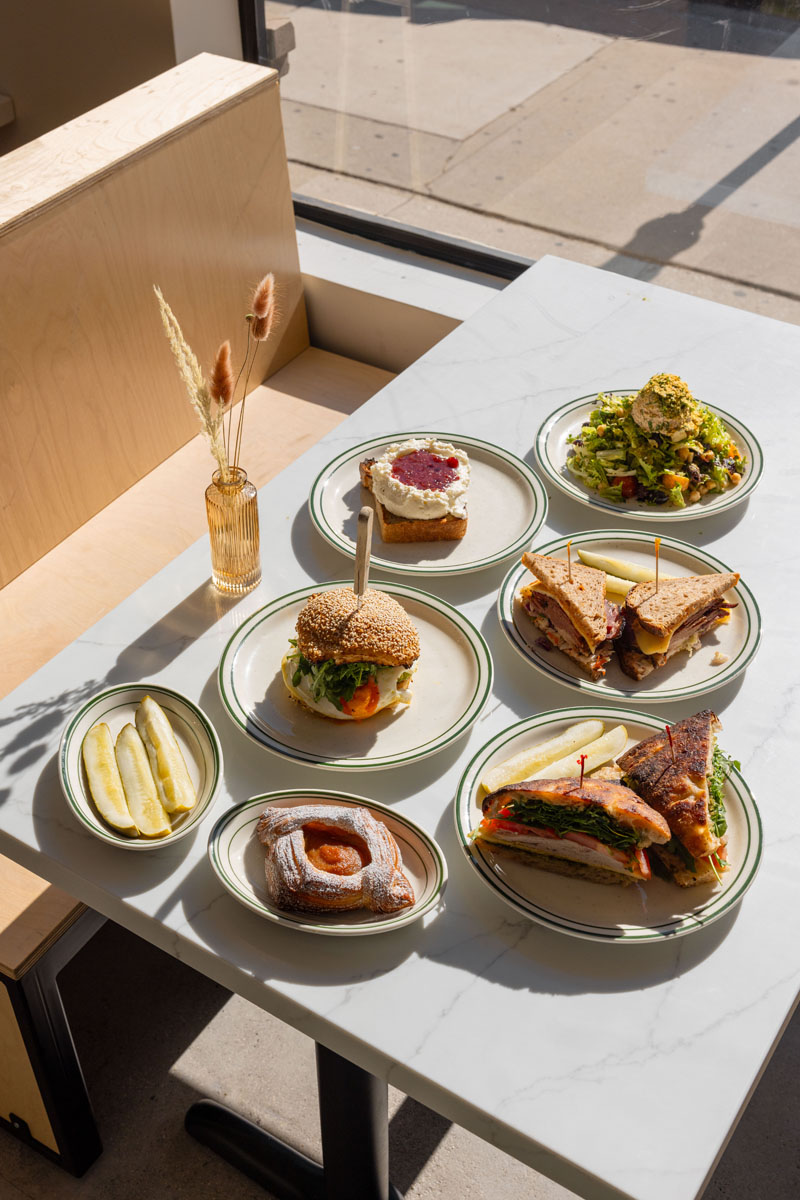 A sunlit table with plates of sandwiches, salad, toast with spread, pickles, and a pastry, set near a window with a small vase of dried flowers.