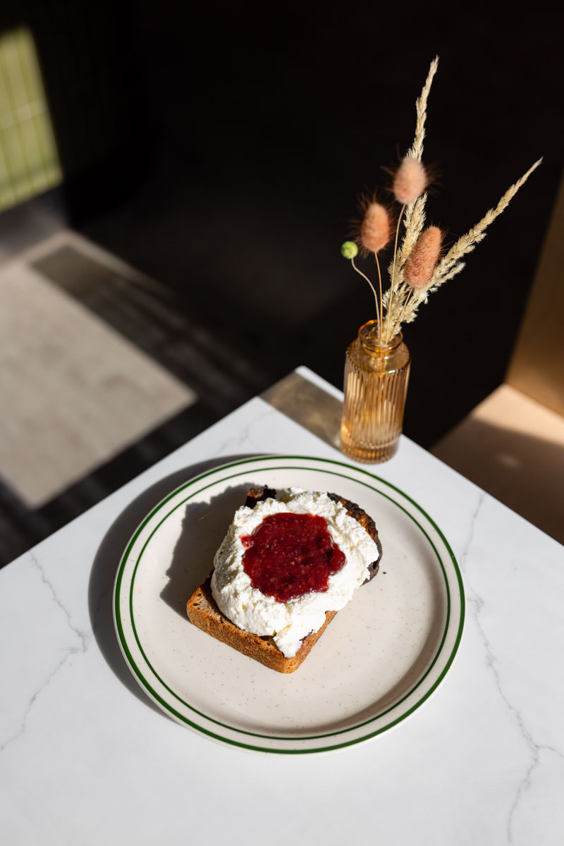 A plate with a slice of toast topped with whipped ricotta and a dollop of berry jam sits on a white table next to a small amber vase holding dried grasses. Sunlight casts shadows across the scene.