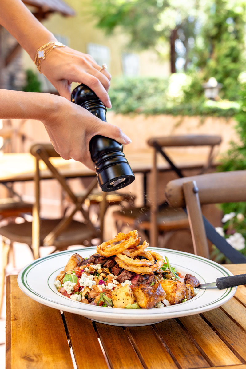A person’s hands grind pepper over a plated salad topped with onion rings and various vegetables, on a wooden outdoor table with chairs and greenery in the background.