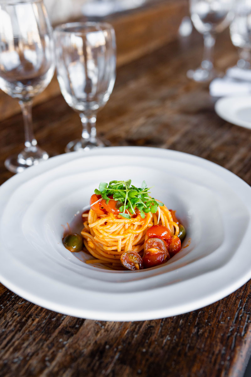 A plate of spaghetti with tomato sauce, cherry tomatoes, olives, and fresh microgreens sits on a rustic wooden table, surrounded by empty wine glasses in the background.