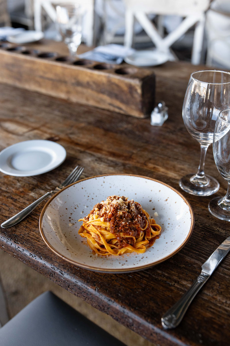 A plate of tagliatelle pasta topped with meat sauce sits on a rustic wooden table, set with cutlery, empty wine glasses, and a small bread plate.