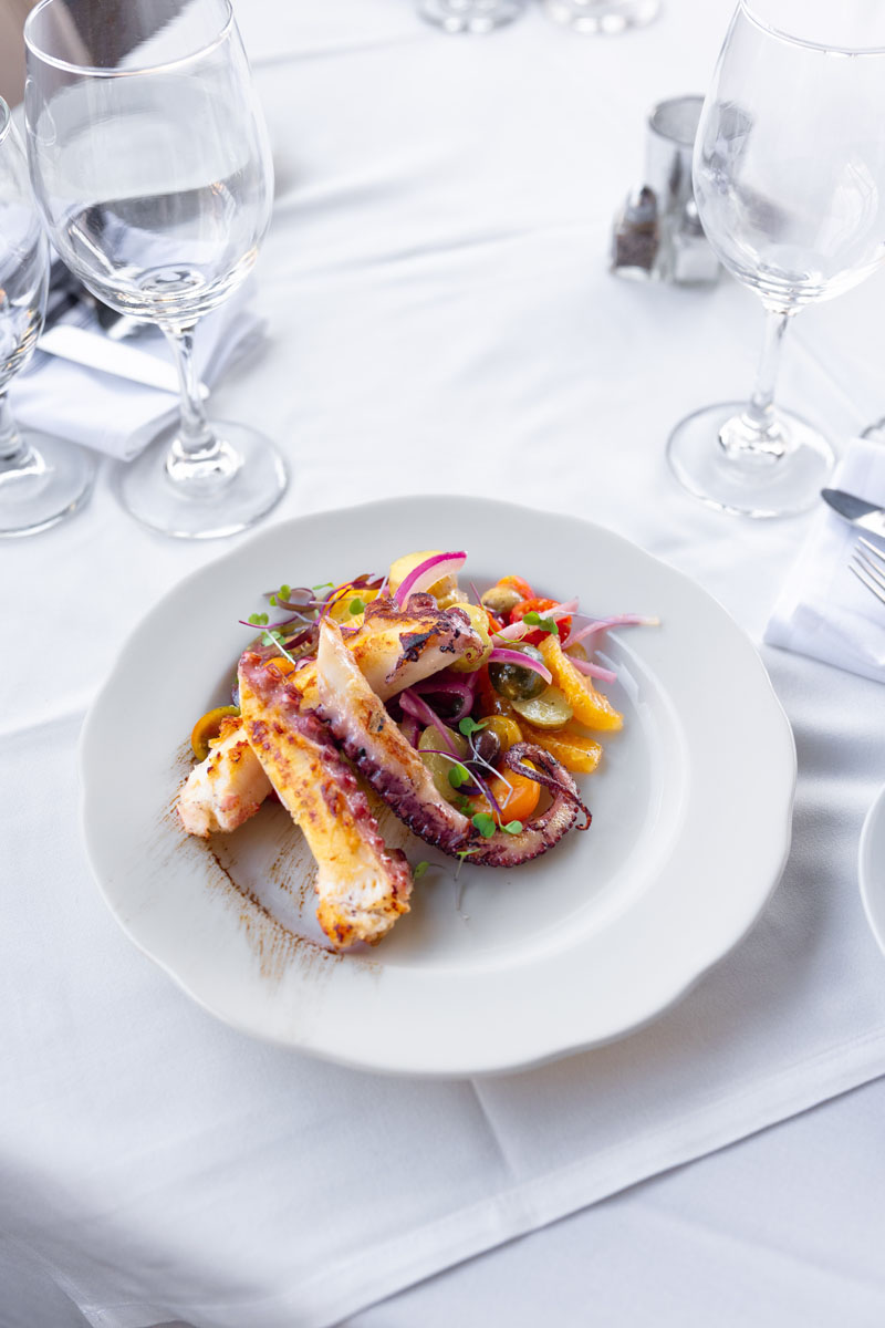 A white plate with grilled octopus, colorful cherry tomatoes, red onion, and microgreens is set on a white tablecloth, surrounded by empty wine glasses and folded napkins.
