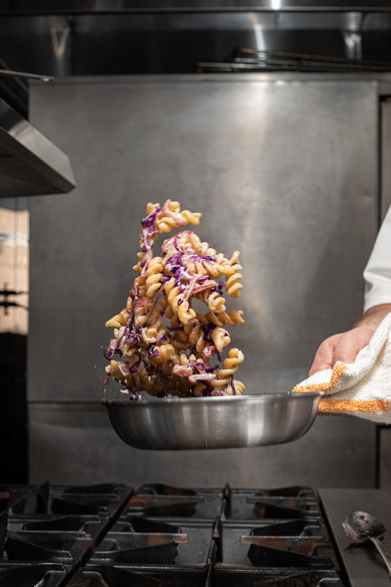 A chef tosses pasta with purple cabbage in a stainless steel pan over a stovetop, capturing the ingredients midair. The chef’s hand holds the pan with a towel in a professional kitchen setting.